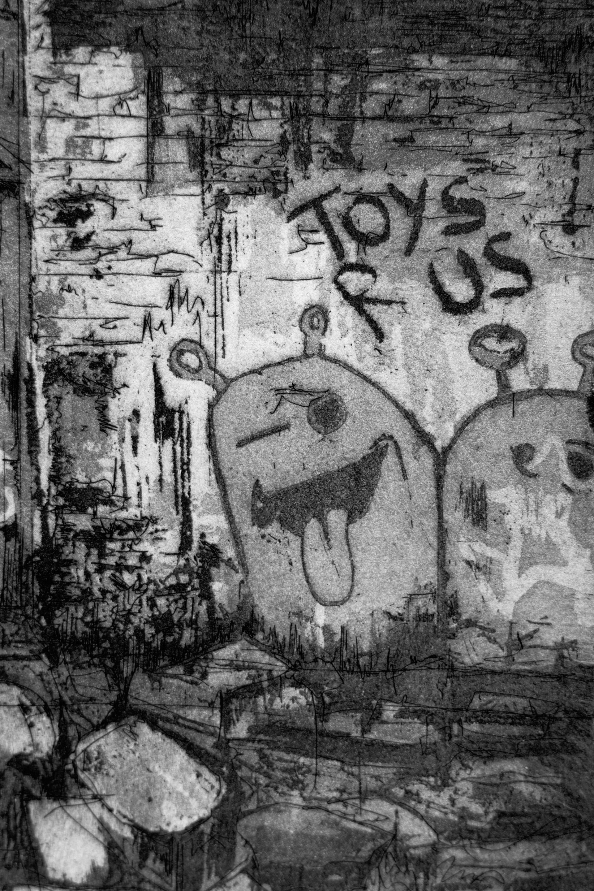 Detailausschnitt der Radierung "The British Industrial Estate" von Jemma Gunning: Nahaufnahme des "Toys R Us"-Graffitis mit gezeichneten Cartoongesichtern auf der Backsteinwand.