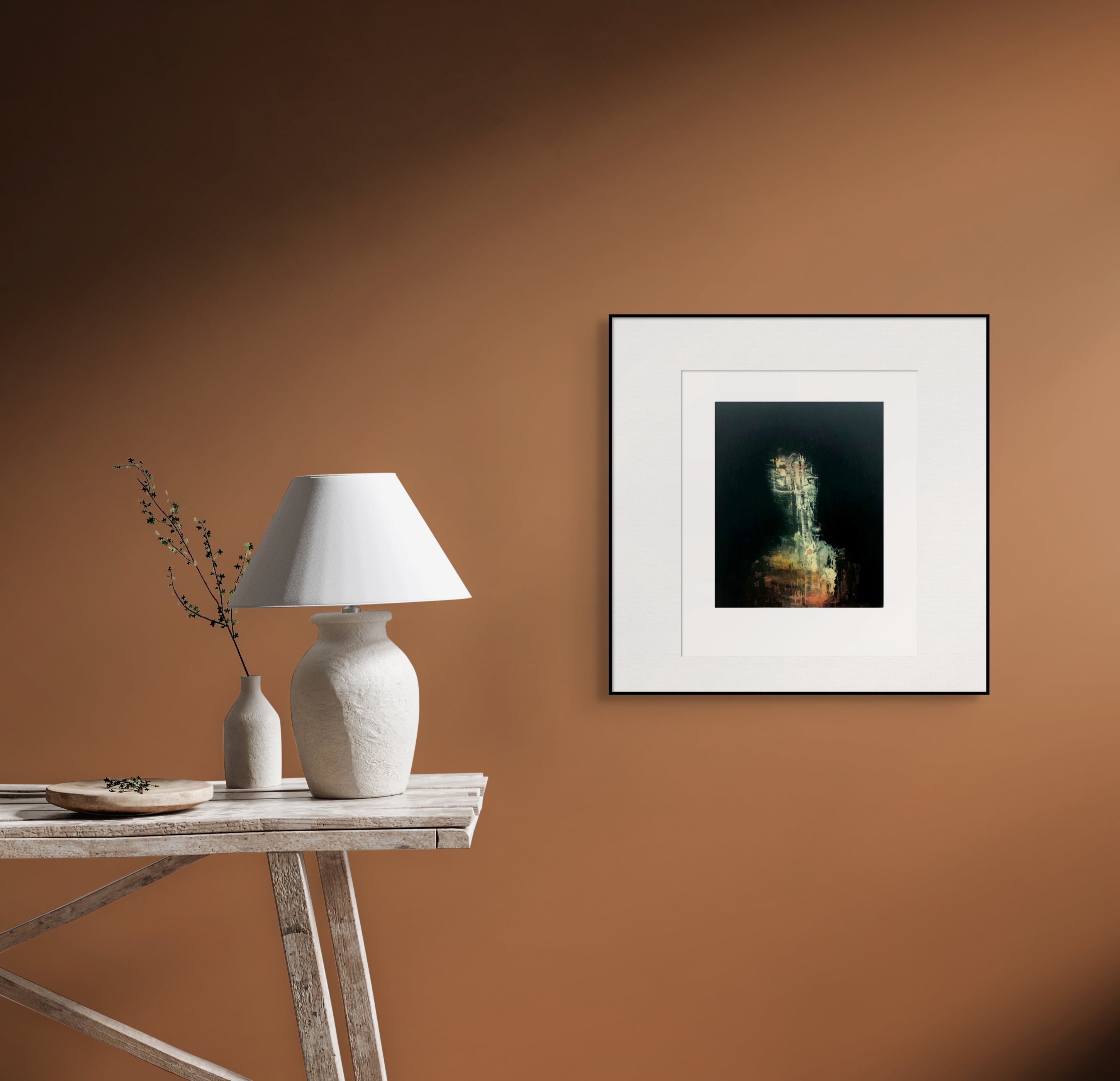 Gerahmte Radierung "Revenant" von Stephen Lawlor – Gerahmtes Bild an warm beleuchteter Terrakottawand neben rustikalem Holztisch mit weißer Lampe.